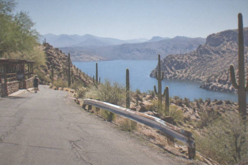 Saguaro cacti and rocky desert slopes along a paved highway leading toward a lake between canyon walls