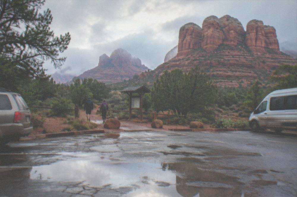 A trailhead parking area with red rock buttes rising behind parked vehicles in morning light