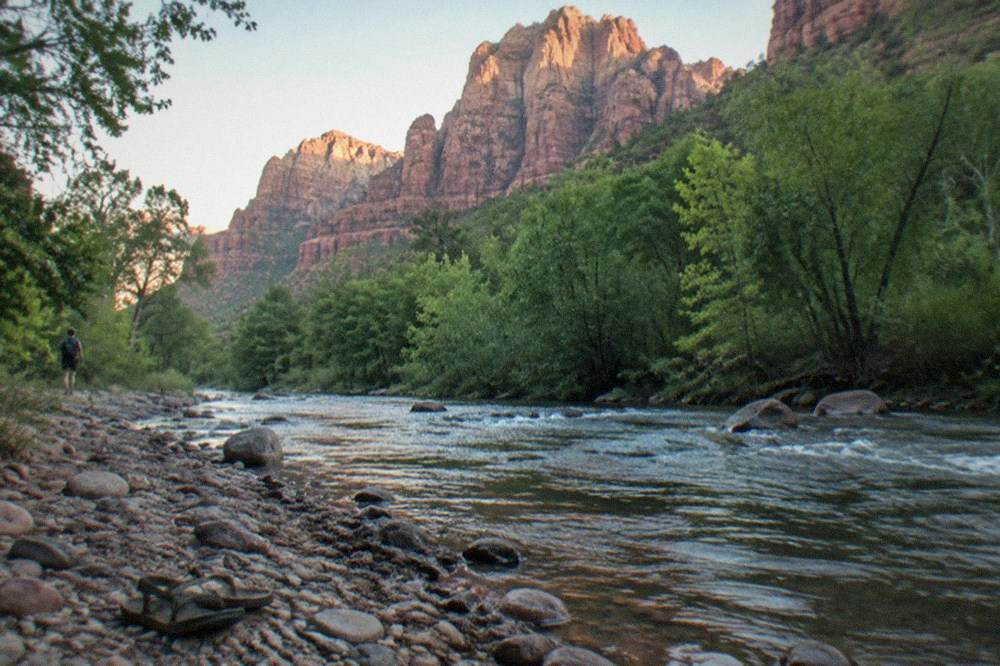 Towering red sandstone buttes above a creek lined with green trees in a canyon valley