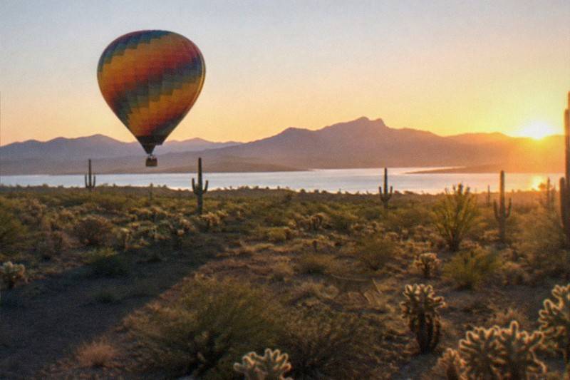 Hot air balloons float above open desert outside Phoenix at sunrise, saguaro cacti scattered across the ground below.