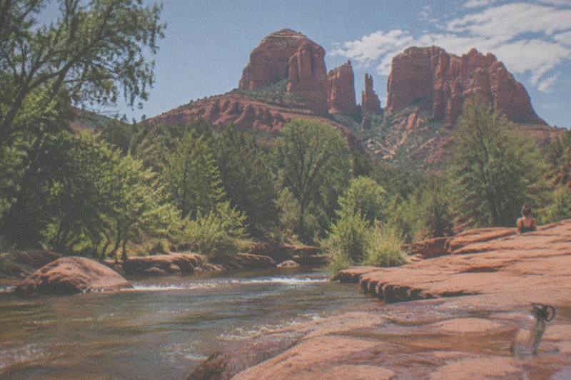 Late sun lights the red rock towers above Oak Creek in Sedona, where green trees gather along the water below the cliffs.