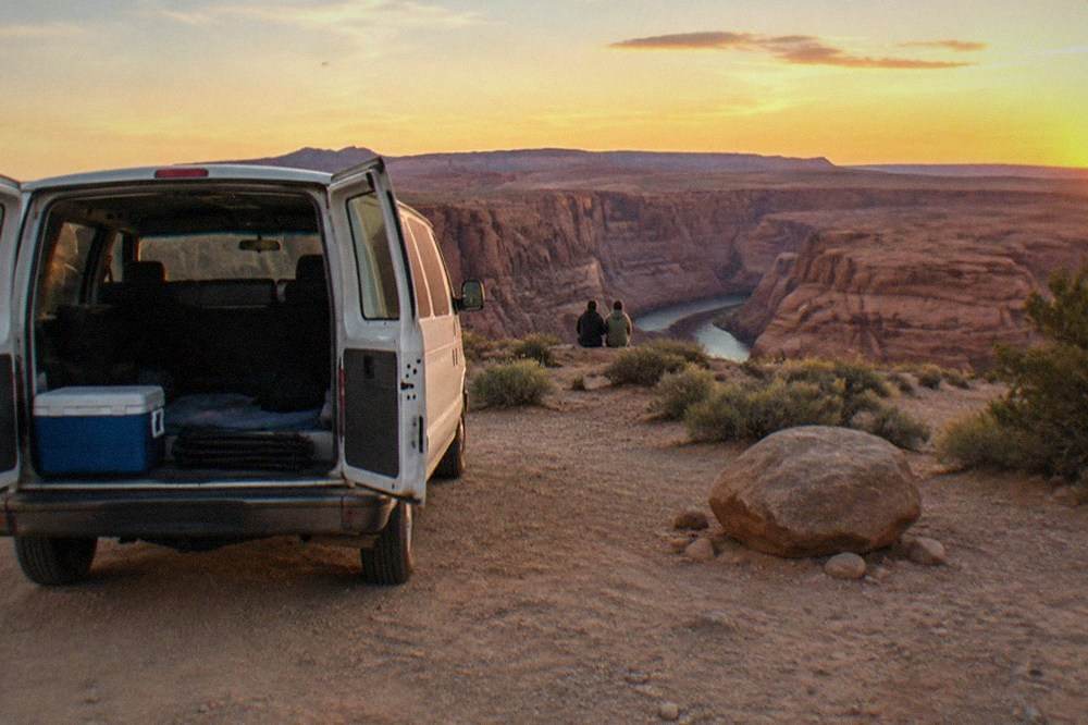 A tour van parked at a desert overlook with red rock mesas in the distance under a wide sky