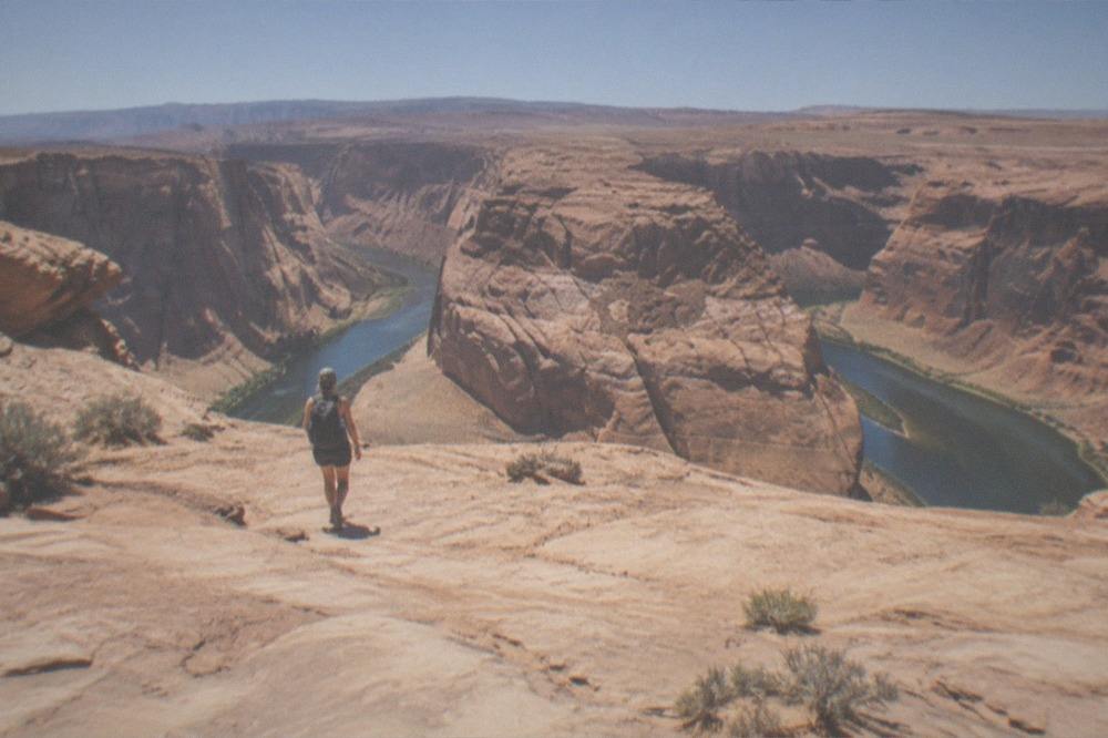A hiker with a daypack walking across open sandstone toward a canyon rim viewpoint