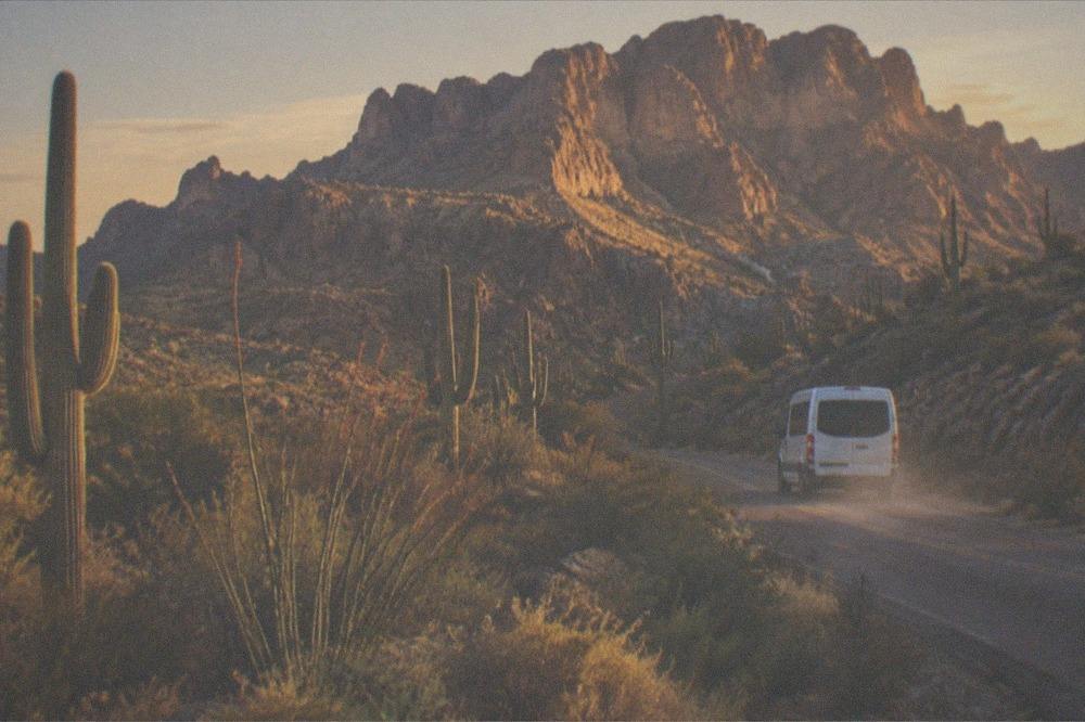 A tour van traveling a winding desert road beside saguaro cacti in golden light