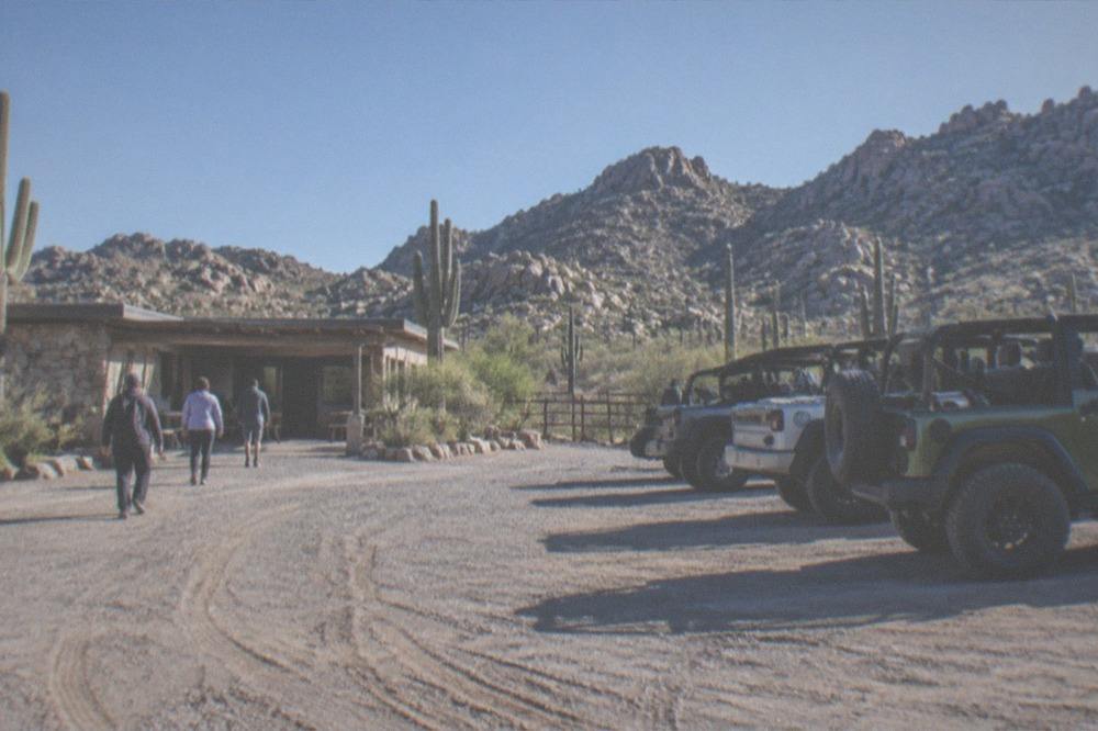 A desert staging area with parked vehicles and a trailhead sign at the base of rocky hills