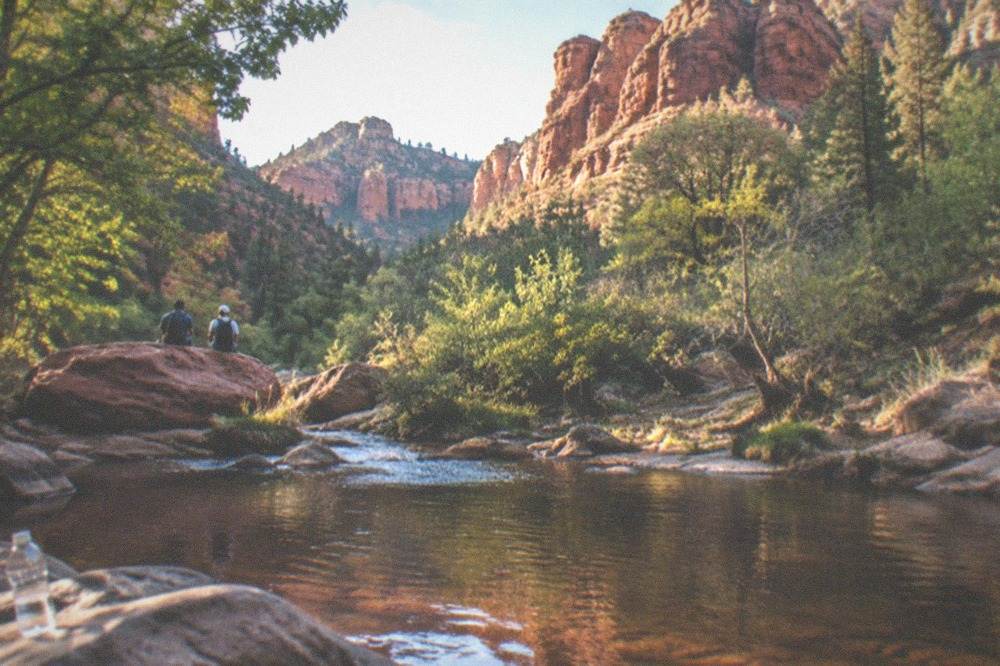 Red rock buttes and forested cliffs rising above a creek corridor in late afternoon light