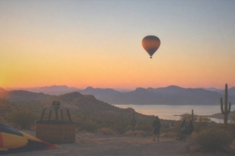A hot air balloon climbing into a pink and orange sunrise sky above distant desert ridgelines
