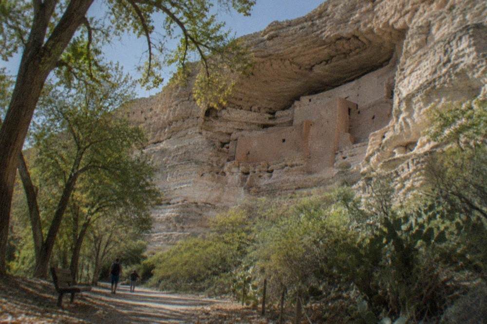 Stone cliff dwelling set into a recessed limestone cliff above a creek lined with cottonwood trees