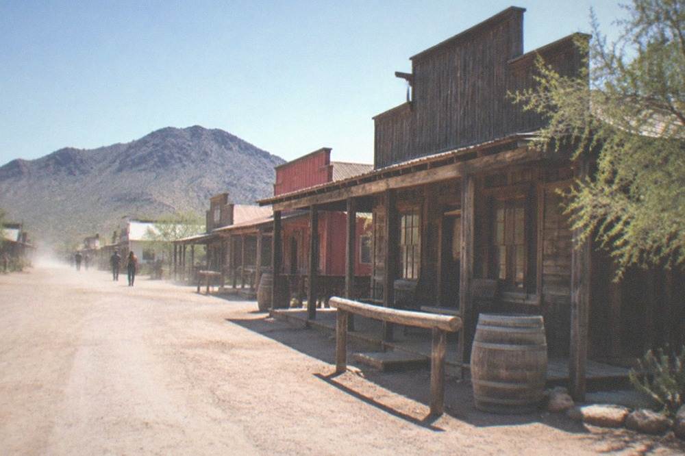 Historic wooden storefronts of an old west town with a dirt street and boardwalk in bright daylight